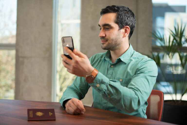 man in green long-sleeved shirt holding his phone up with passport laying on the table